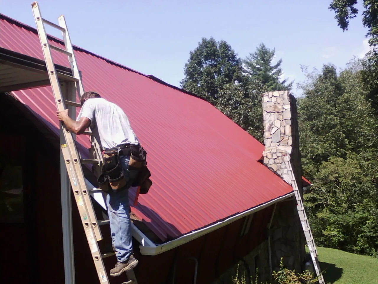 Roofer on a ladder beside a red metal roof and stone chimney — Soffit & Fascia Repair by Roofing Brothers in Cressey
